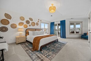 Une chambre à coucher comprenant un lit, des tables de chevet, une lampe, un tapis à motifs, un luminaire au plafond, une fenêtre avec des rideaux et une vue sur l’extérieur.