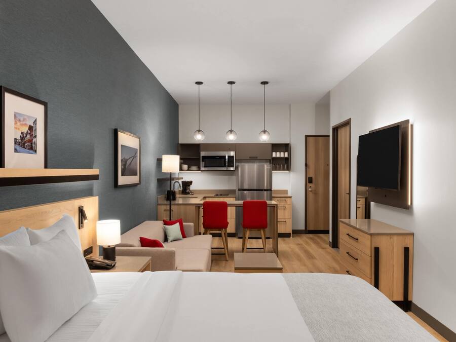 A modern hotel room with a bed, a desk, a kitchenette, and a TV.