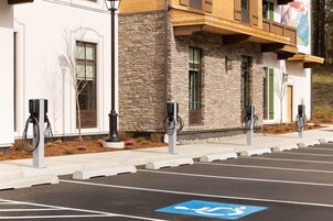 Un estacionamiento con estaciones de carga para vehículos eléctricos, un espacio reservado para personas con movilidad reducida y un edificio con fachada de piedra y madera.