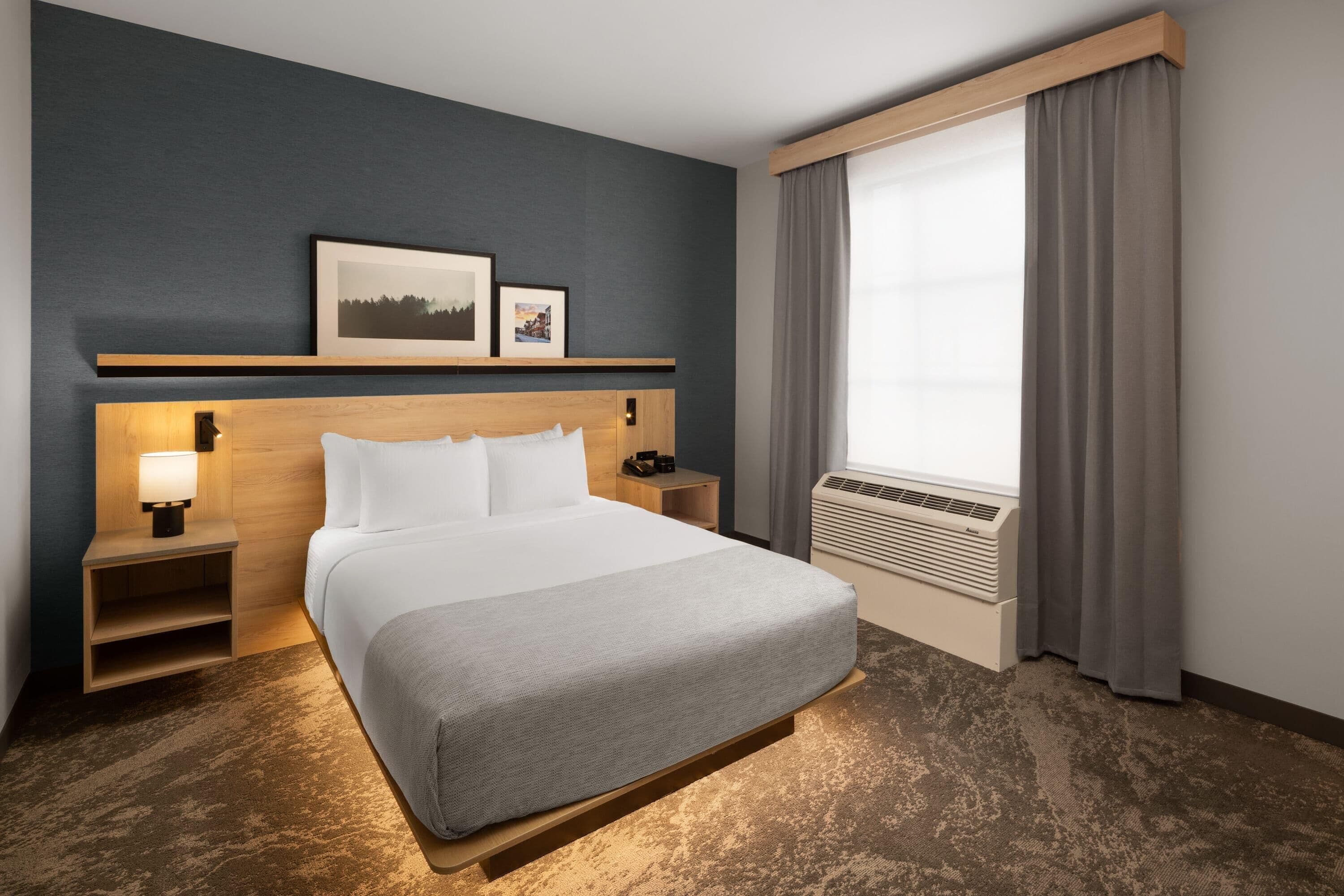 Ett hotellrum med en säng, sängbord, en vägghylla med bilder, ett fönster med gardiner och en luftkonditioneringsanläggning.