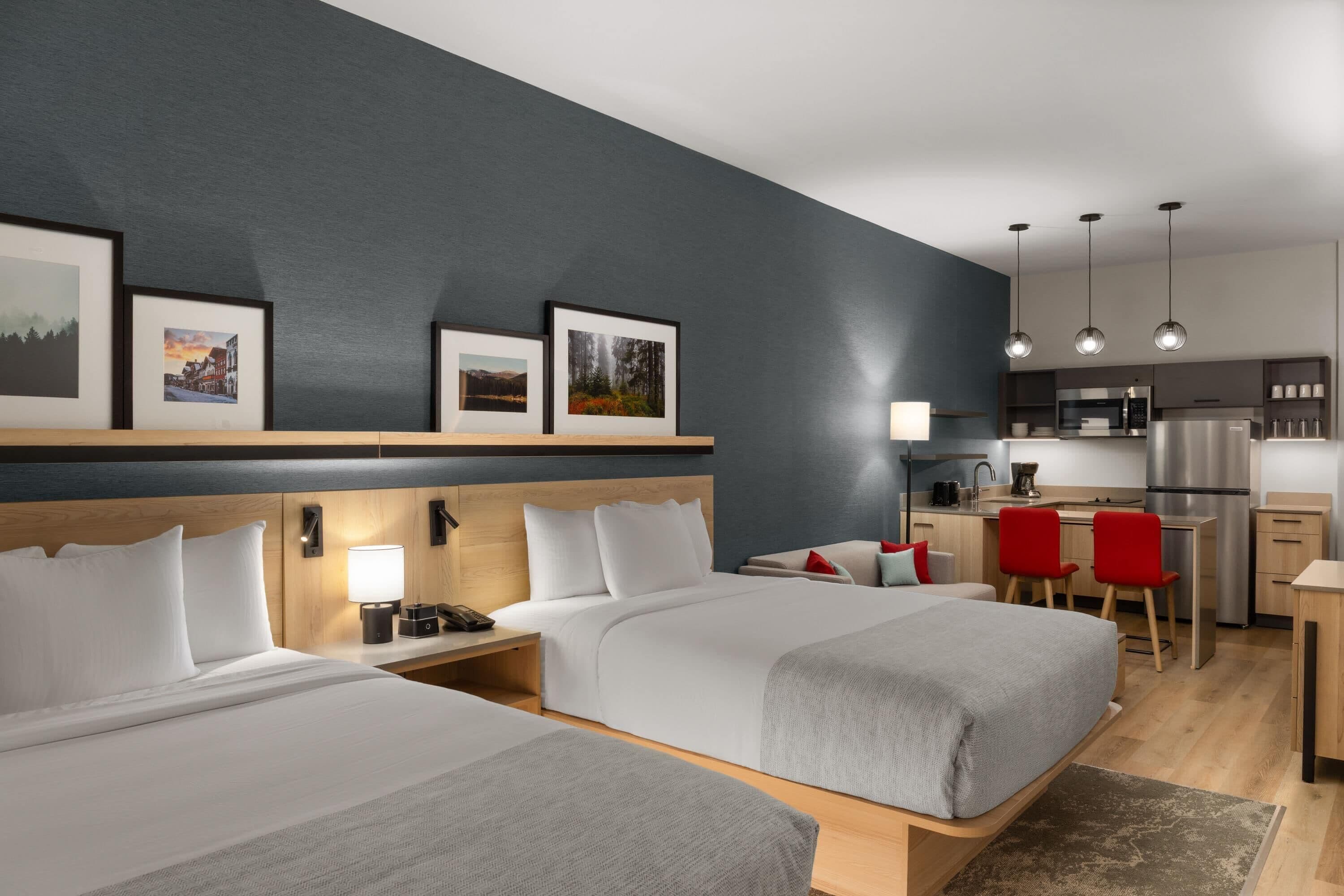 Una habitación de hotel moderna con dos camas, un escritorio, kitchenette y cuadros en la pared.