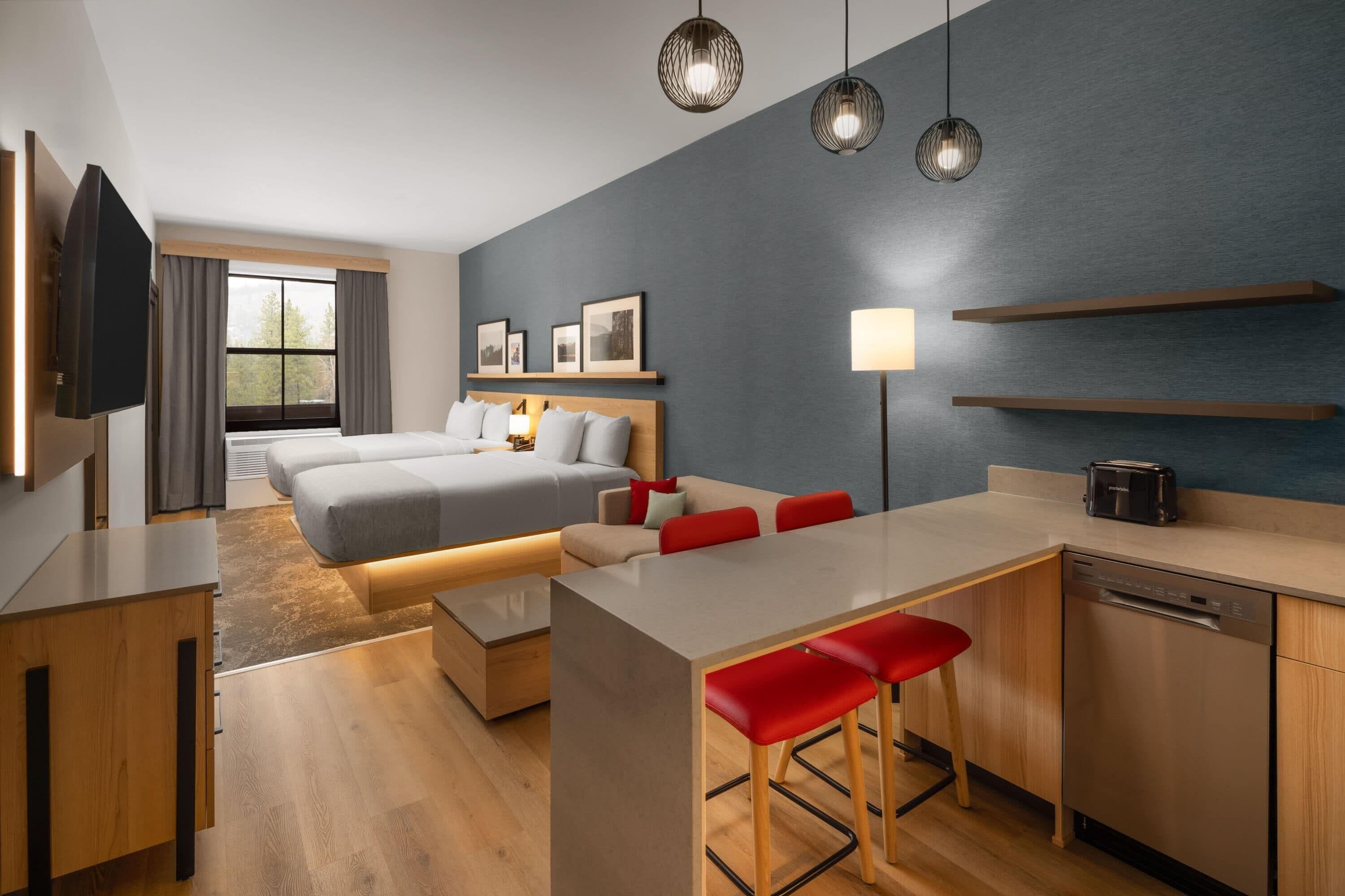 A modern hotel room with a bed, a desk, a kitchen area, and a dining space.