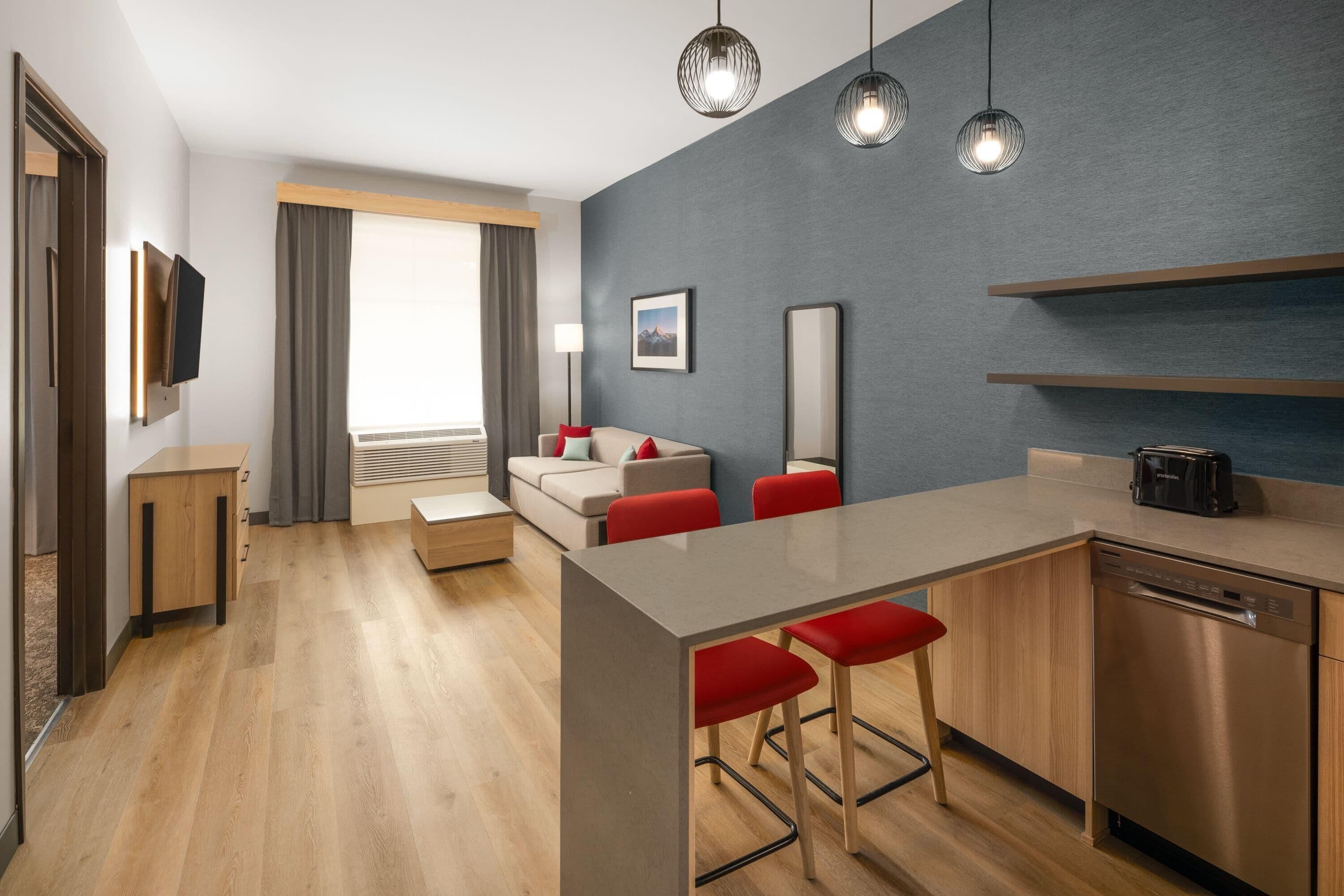 A modern hotel room with a wooden floor, a kitchenette, a sofa with red cushions, a TV mounted on the wall, and a large window with curtains.