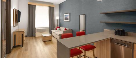 A modern hotel room with a wooden floor, a kitchenette, a sofa with red cushions, a TV mounted on the wall, and a large window with curtains.