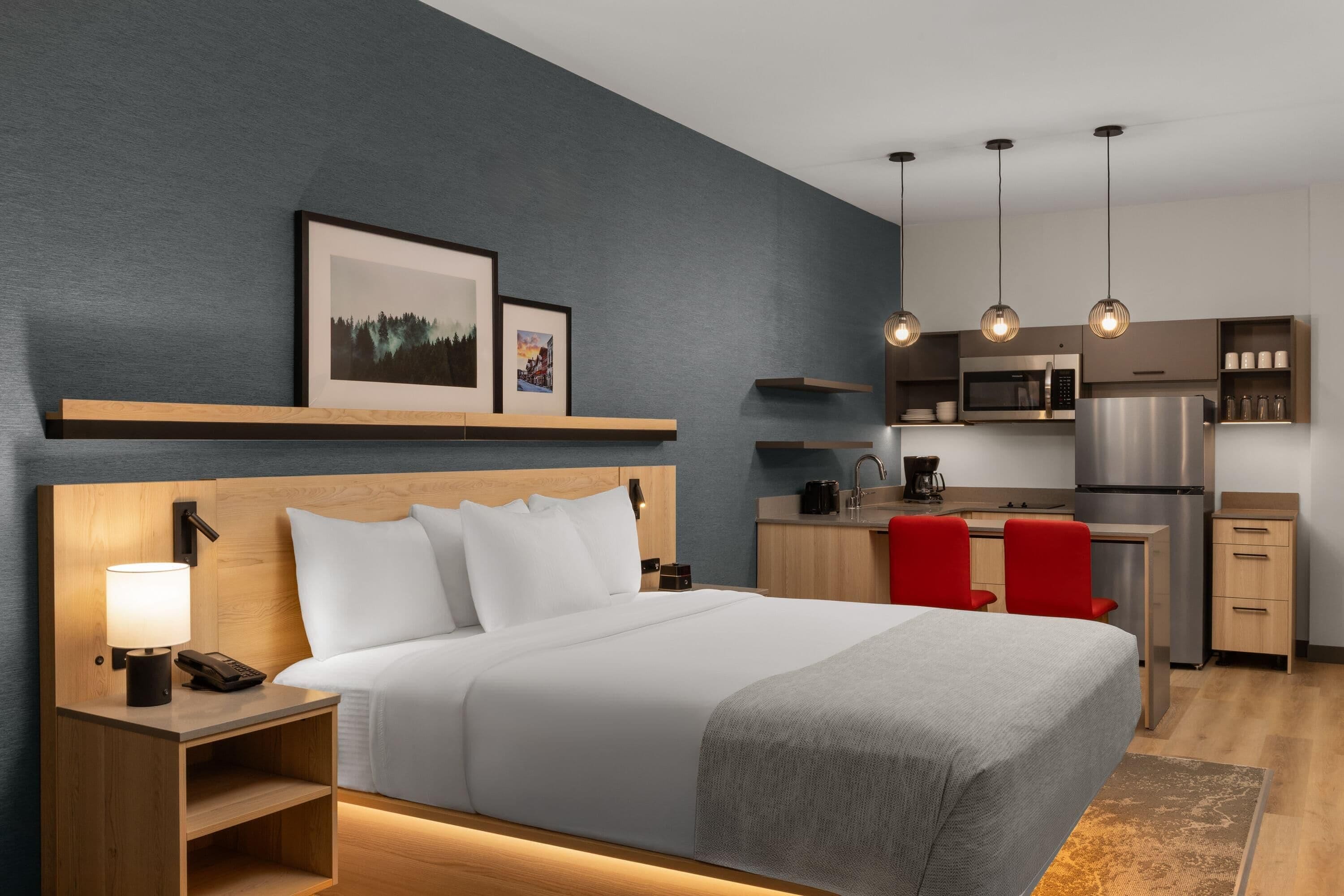 Une chambre d’hôtel comprenant un lit, une table de chevet, une lampe et un coin cuisine équipé d’un four à micro-ondes, d’un réfrigérateur et de chaises rouges.
