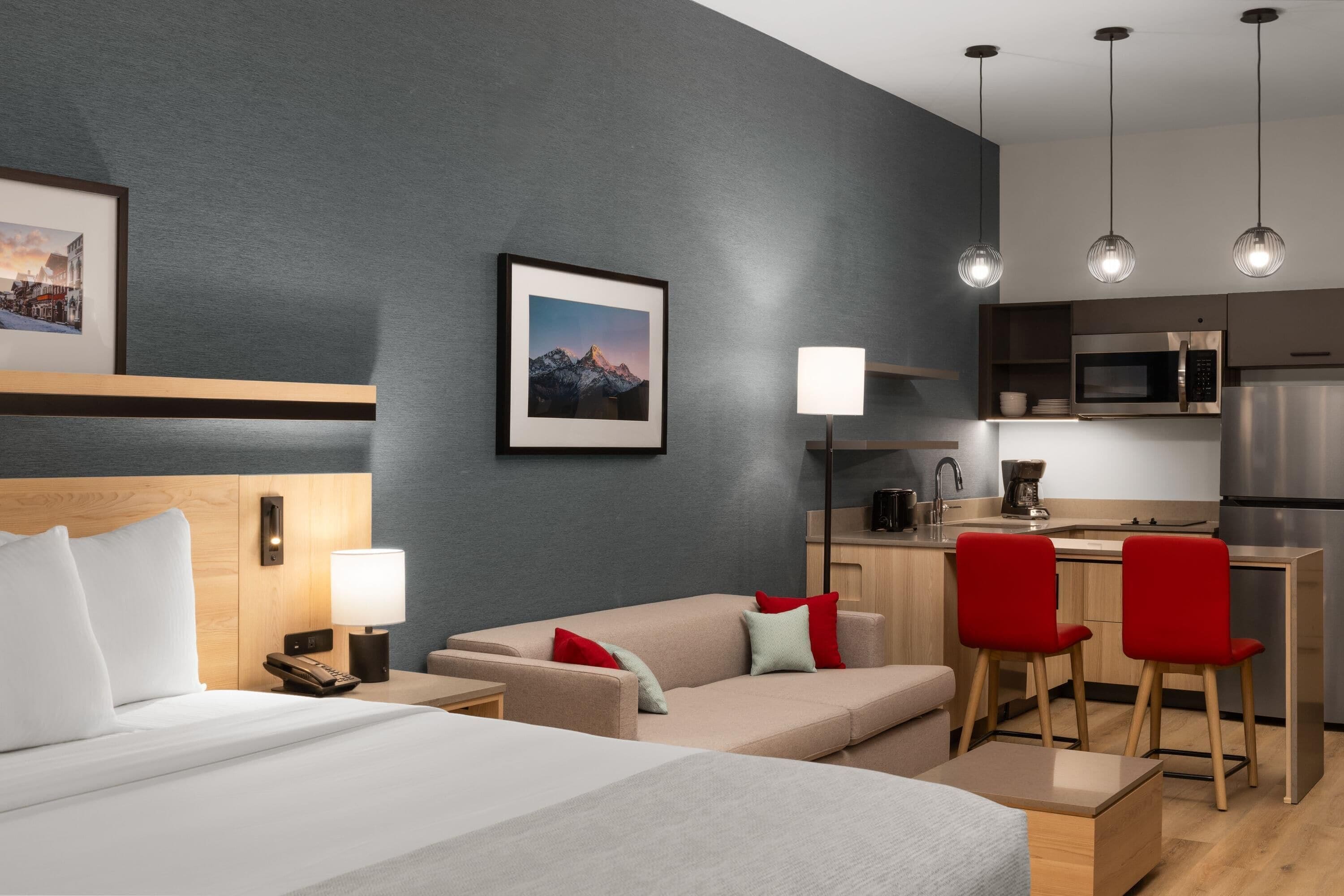 Une chambre d’hôtel moderne équipée d’un lit, d’un canapé, d’un bureau avec des chaises, d’une kitchenette et d’un tableau encadré au mur.