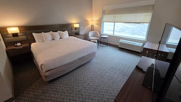 A hotel room with a large bed, bedside tables with lamps, a chair, a small table, a TV, and a window with blinds.