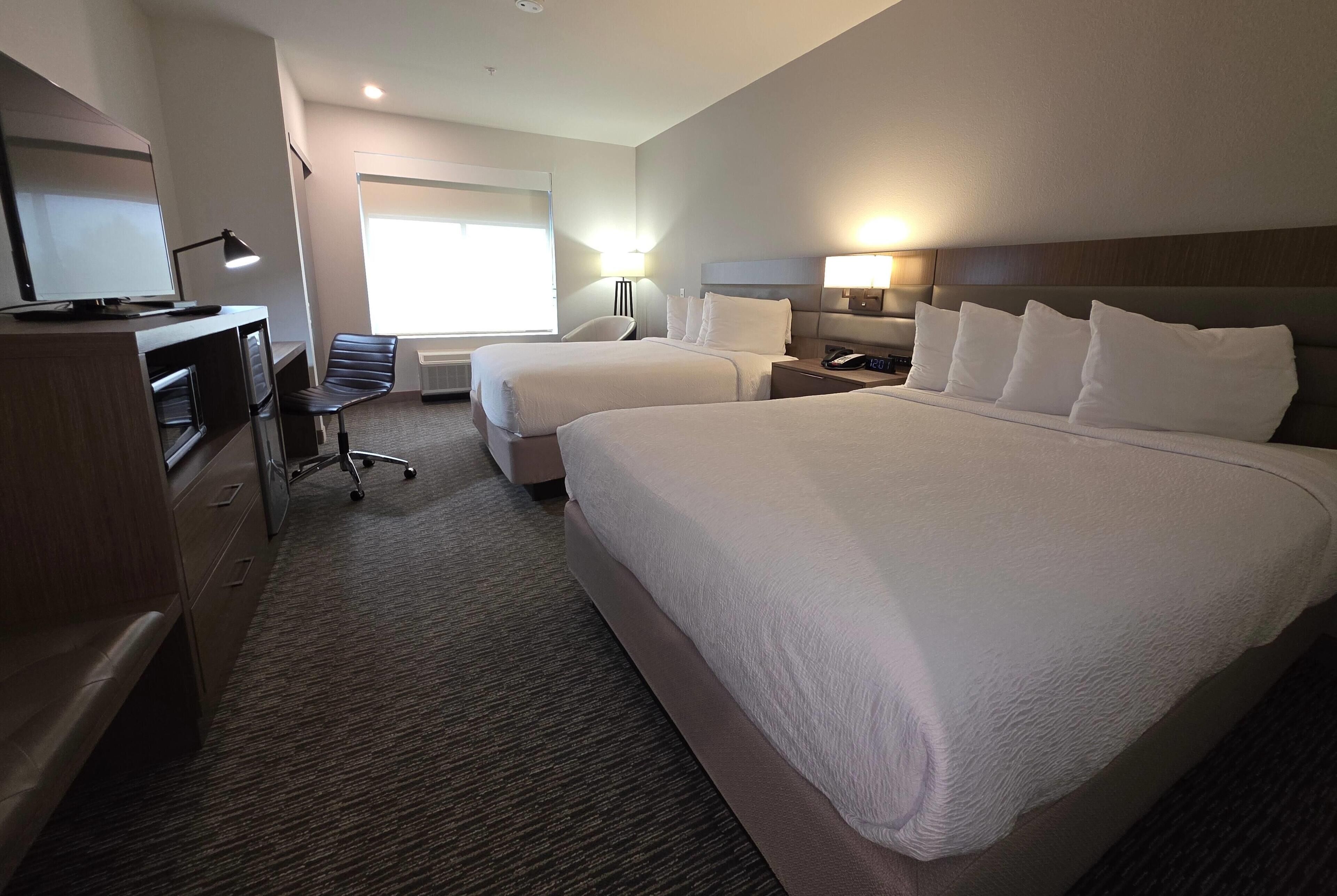 A hotel room with two beds, a desk, a chair, a TV, and a window with natural light.
