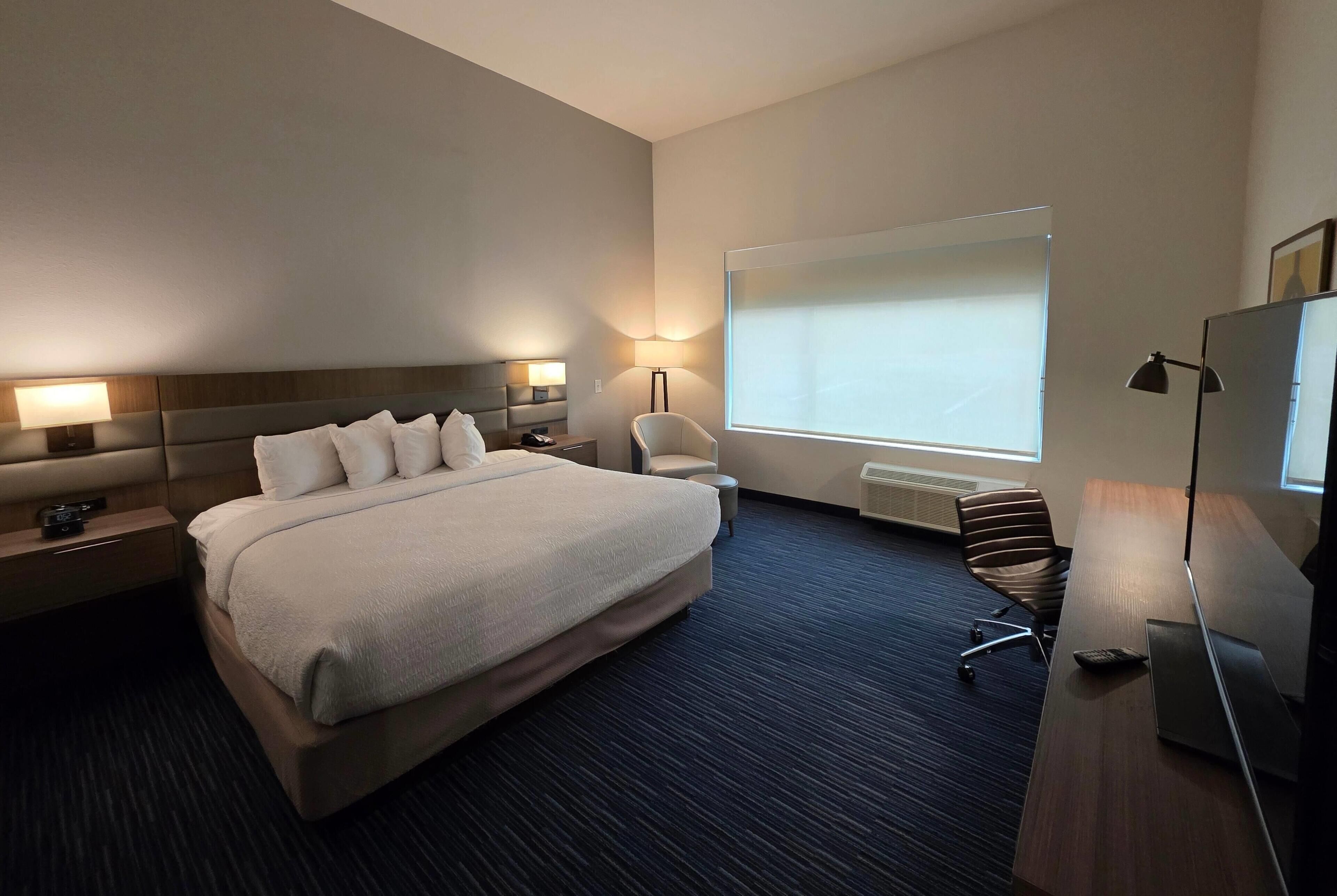 A hotel room with a large bed, a desk with a laptop, a chair, a bedside table with a lamp, and a large window with blinds.