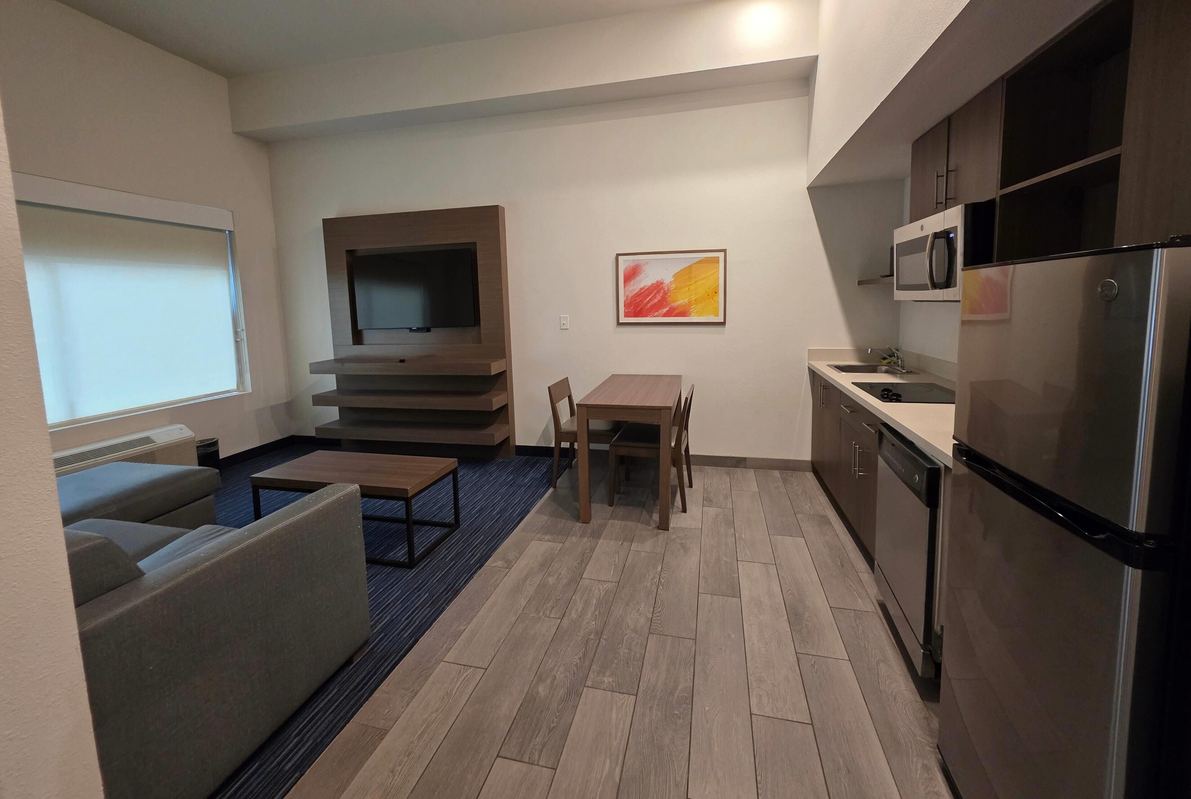 A modern kitchen with a refrigerator, microwave, and dishwasher. There is a dining table with chairs and a TV mounted on the wall.