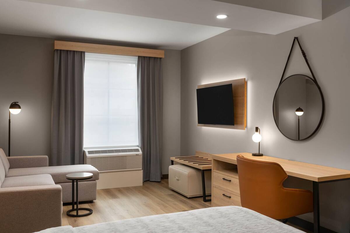 Habitació d'hotel moderna amb un sofà, una taula rodona petita, un escriptori amb una cadira, un televisor, un mirall i una finestra amb cortines.