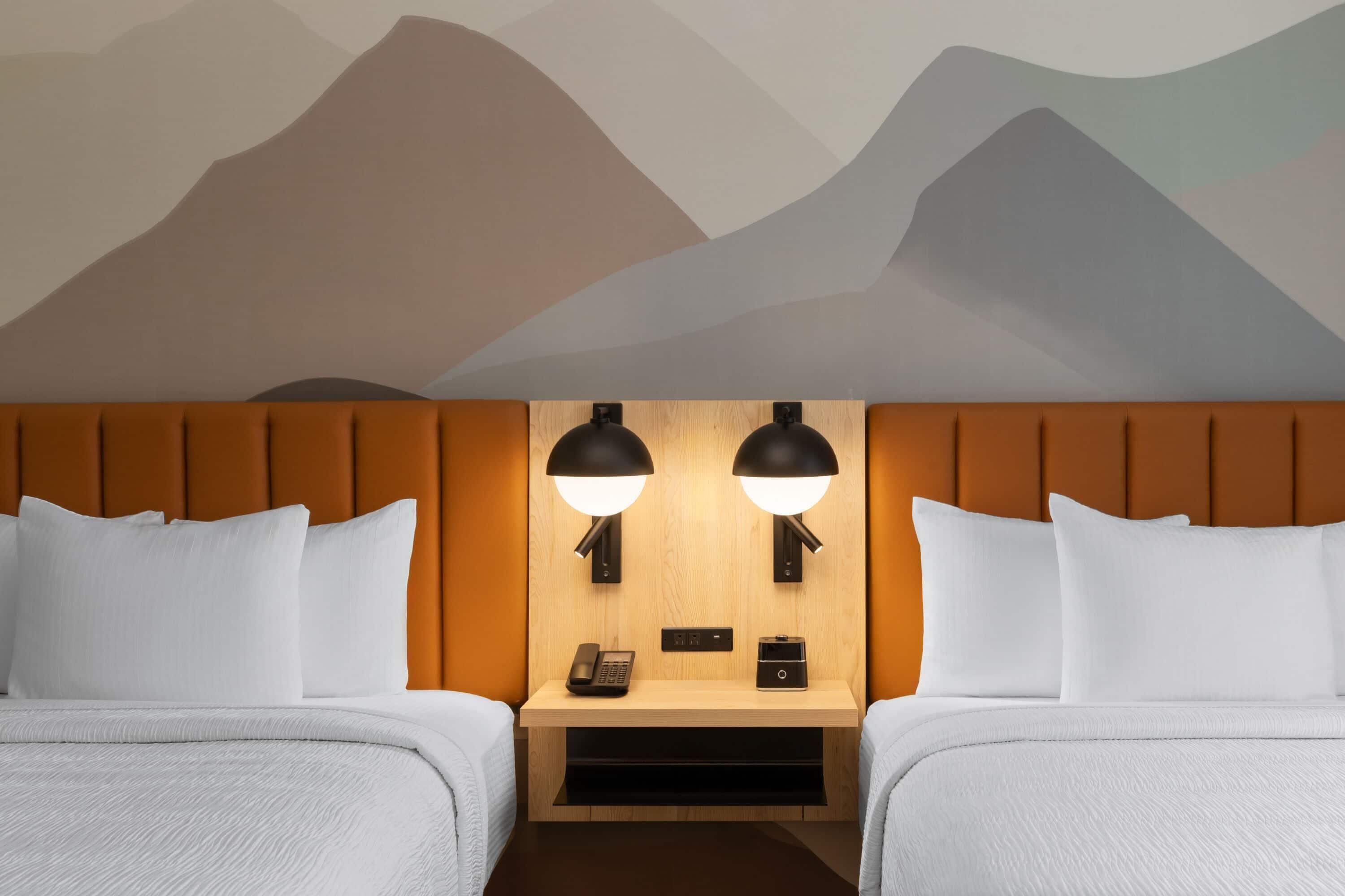 Habitación de hotel con dos camas, cabecero de madera, mesita de noche con teléfono y lámpara, y una pared decorada con un diseño de montaña.