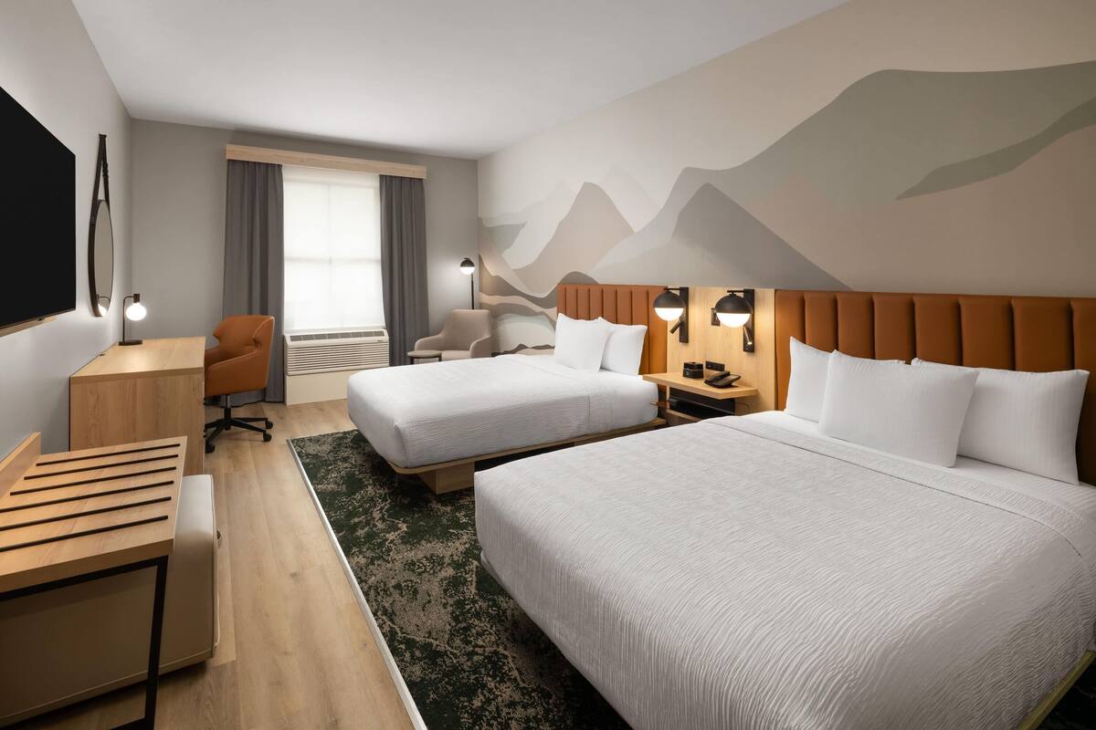 Habitació d'hotel amb dos llits, un escriptori, una cadira, un televisor i una paret amb un disseny de muntanya.