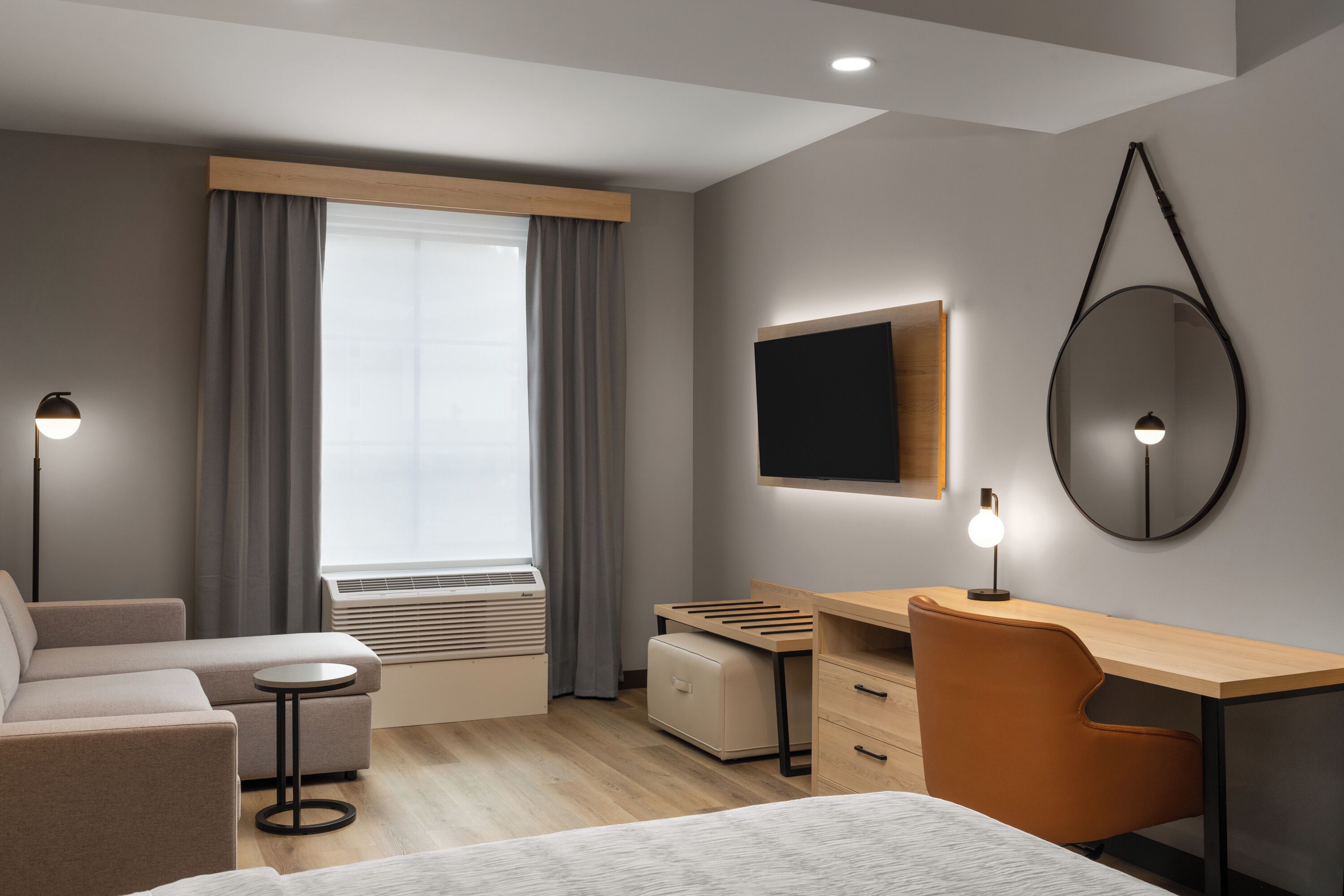 Una habitación de hotel moderna con sofá, escritorio, silla, cama, televisor, espejo y ventana con cortinas.
