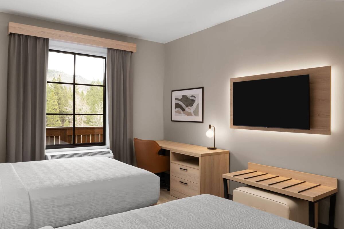 Habitació d'hotel amb dos llits, un escriptori, una cadira, un televisor i una finestra amb cortines.