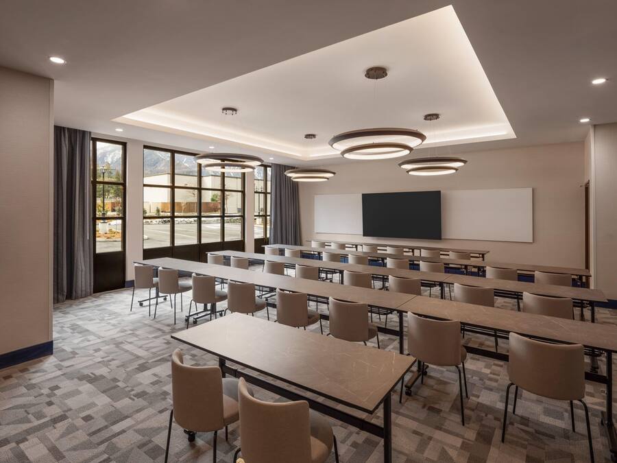 A conference room with long tables, chairs, a projector screen, and large windows with curtains.