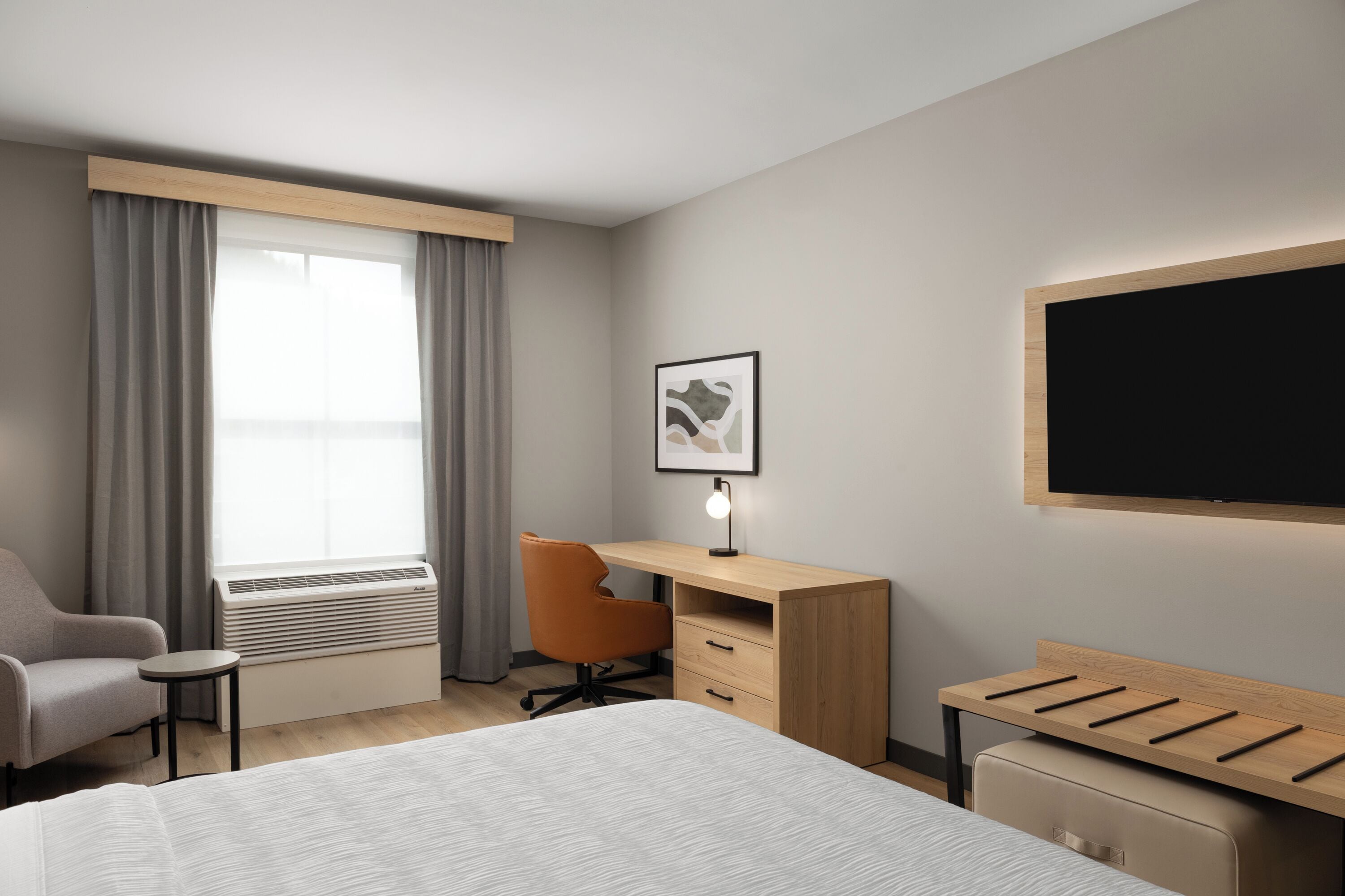 Habitación de hotel con cama, escritorio, silla, radiador, ventana con cortinas, un cuadro en la pared y televisor de pantalla plana montado en la pared.