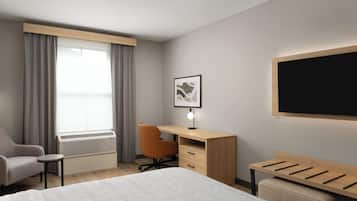 Habitación de hotel con cama, escritorio, silla, radiador, ventana con cortinas, un cuadro en la pared y televisor de pantalla plana montado en la pared.