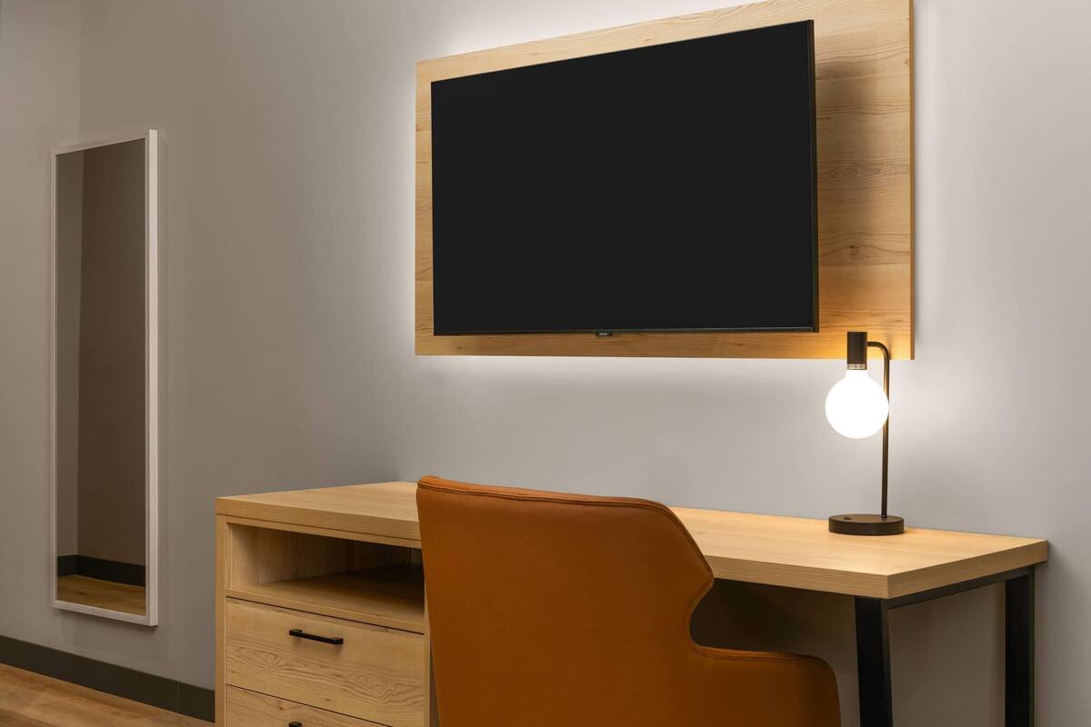 Habitació moderna amb escriptori de fusta, una cadira, televisió penjada a la paret i làmpada de paret.