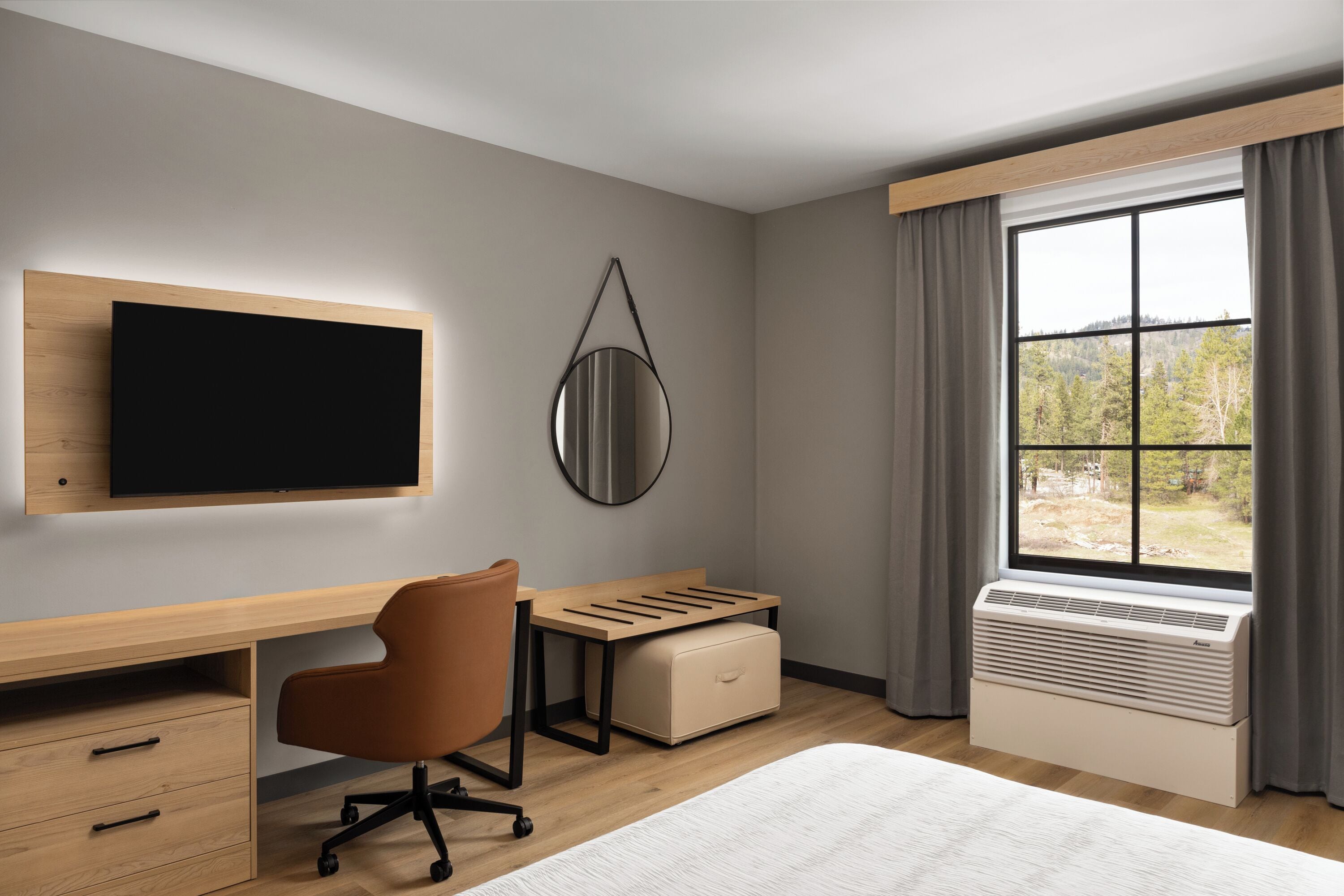 A modern hotel room with a flat-screen TV, a desk with a chair, a window with curtains, and an air conditioning unit.