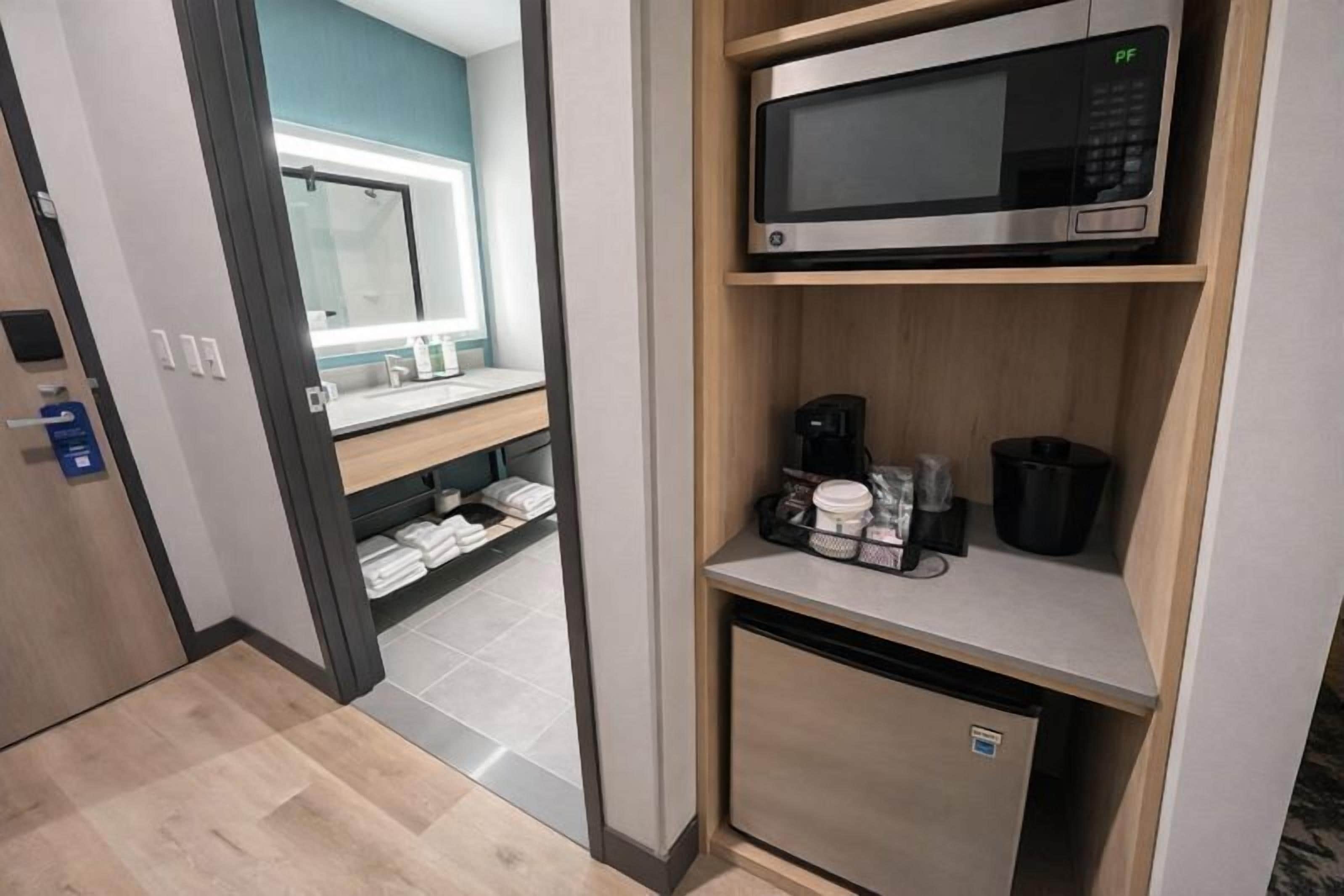 Una habitación de hotel con microondas en un estante, cafetera y un refrigerador pequeño.