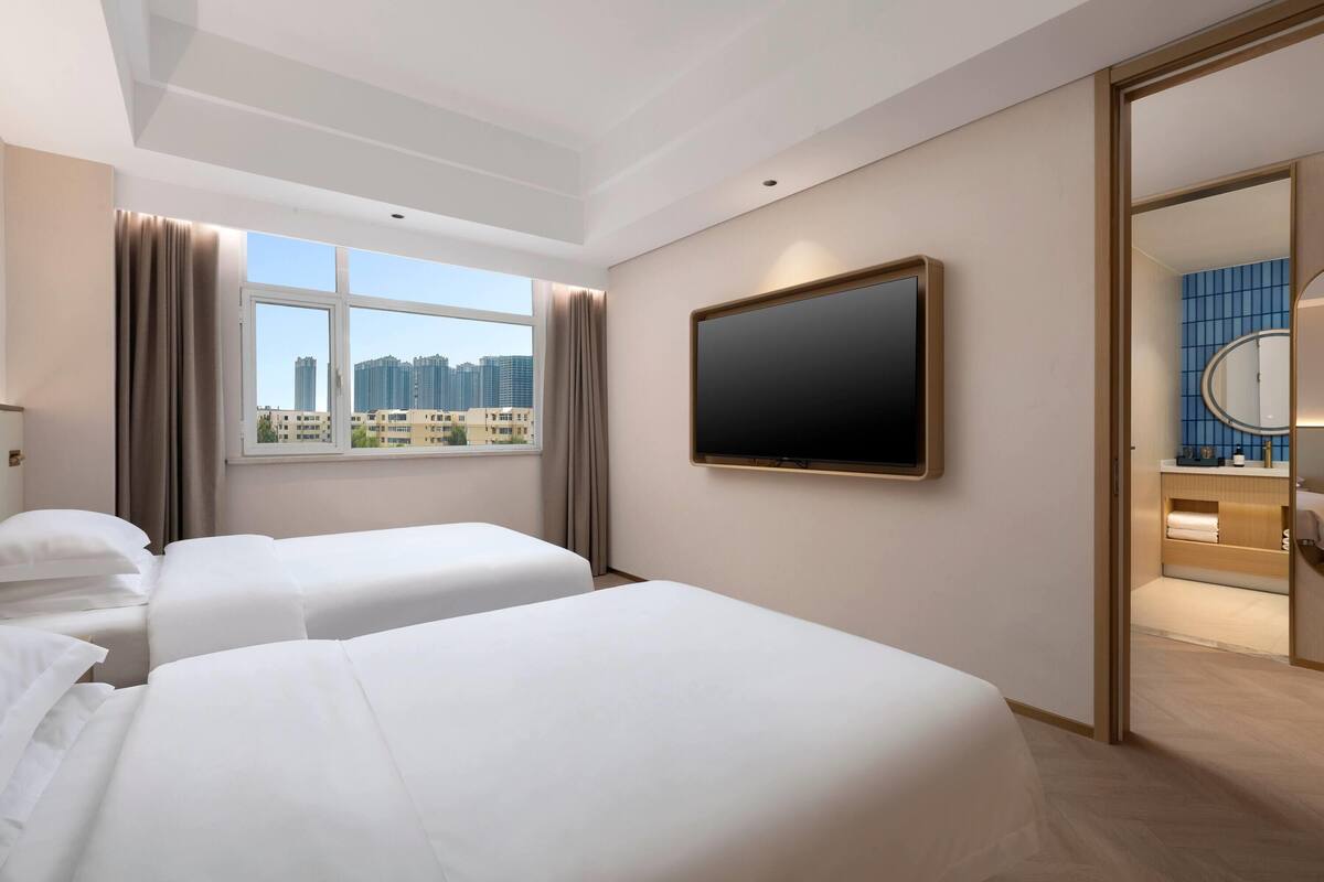 Camera d'albergo con due letti, una grande TV, una finestra con tende e vista su edifici alti.