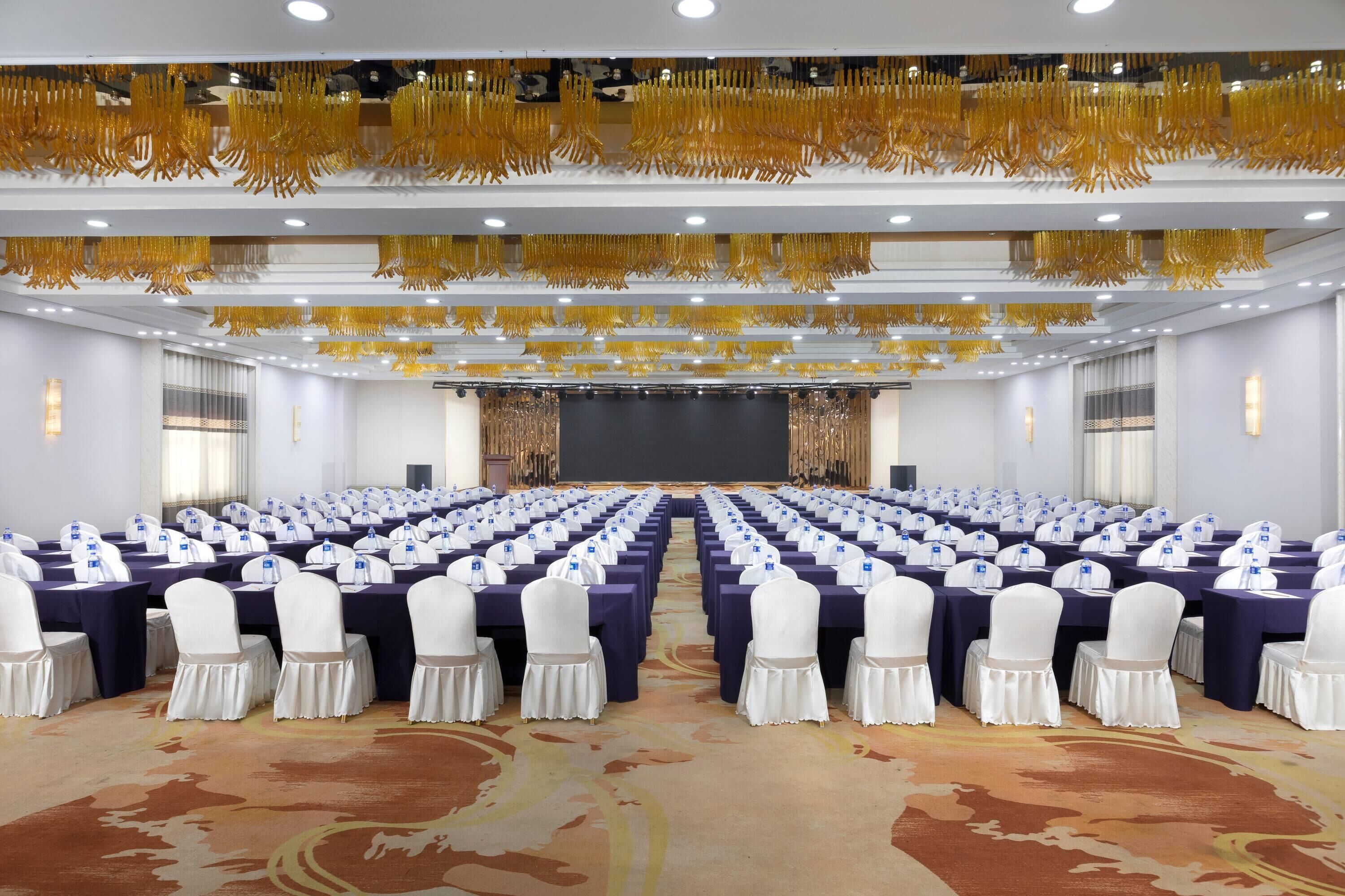 Un salón de conferencias con filas de mesas, sillas blancas y un escenario al frente.