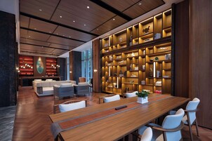 Um hall de hotel moderno com uma mesa de recepção de madeira, sofás brancos, uma longa mesa de madeira com cadeiras e uma grande estante cheia de livros e objetos decorativos.