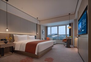 A modern hotel room with a large bed, a desk, a chair, a TV, and a large window with a city view.