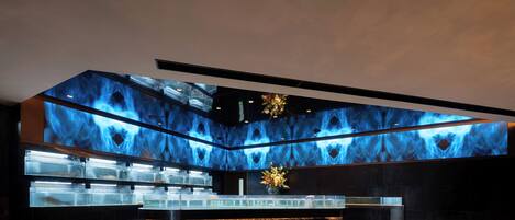 Un interno moderno con un'area bar centrale, un soffitto con motivo luminoso blu e vista sulla città attraverso ampie finestre.