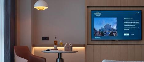A modern hotel room with a brown leather chair, a grey sofa, a small table with a bottle and glass, and a TV displaying a cityscape.
