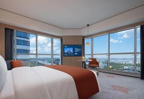 Habitación de hotel moderna con una cama grande, un televisor montado en la pared, una pequeña mesa con una silla y vistas a la ciudad a través de amplias ventanas.