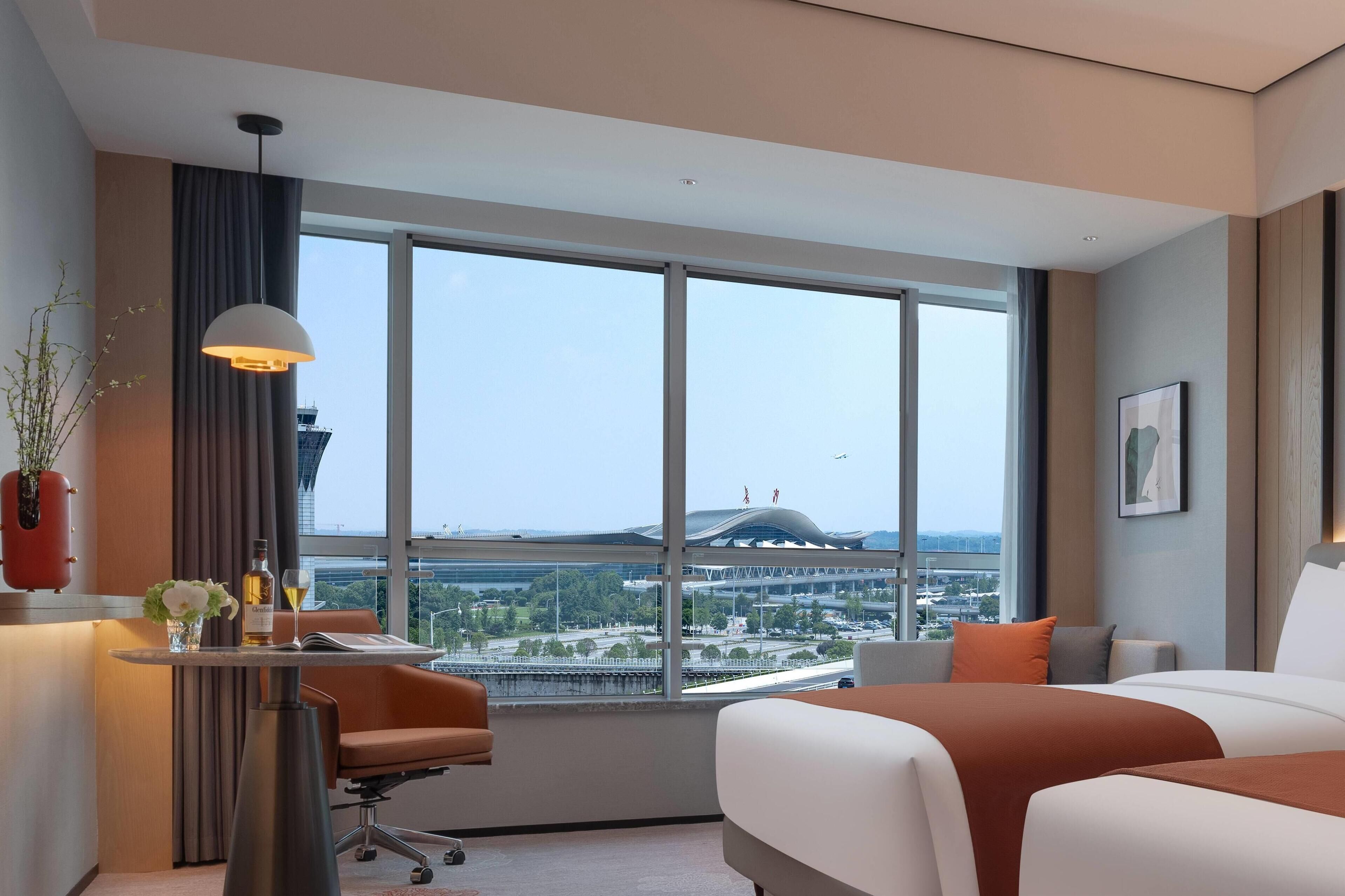 A hotel room with a large window, a bed, a desk with a lamp, and a view of a cityscape.