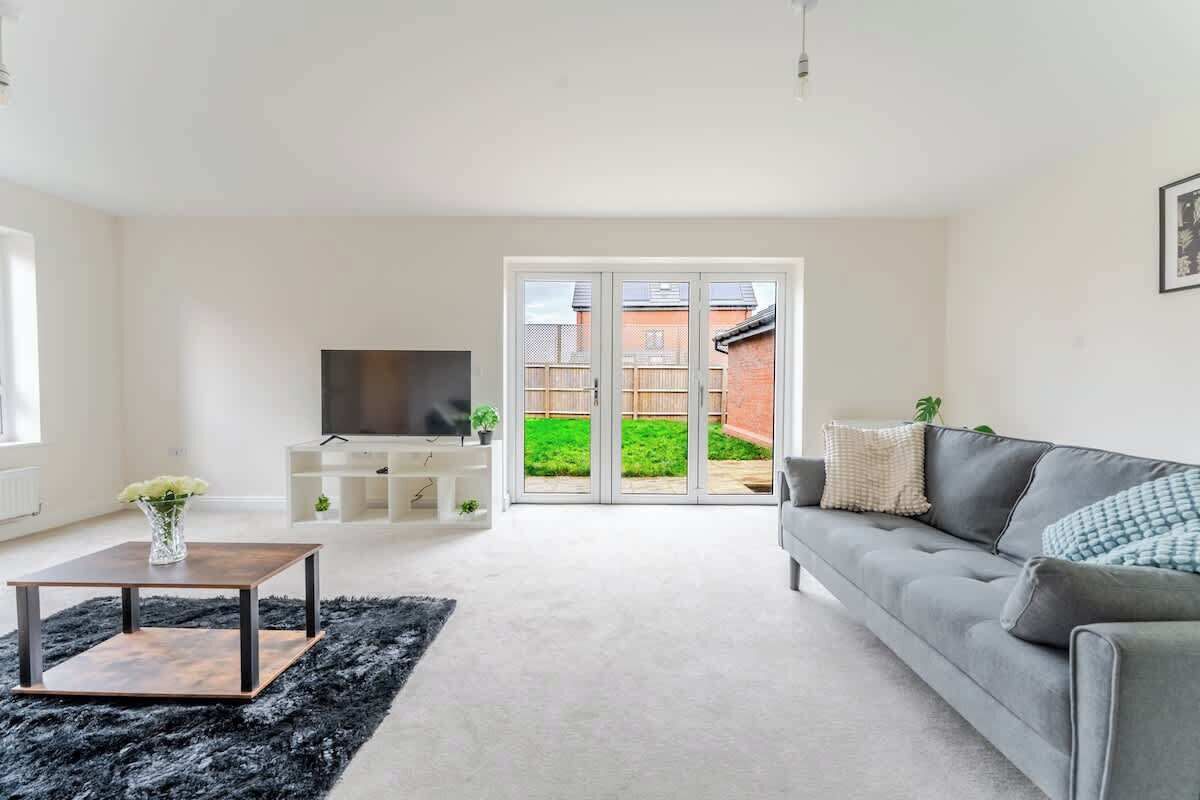 A modern living room with a grey sofa, a TV on a stand, a black rug, and a sliding glass door leading to a garden.