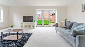 A modern living room with a grey sofa, a TV on a stand, a black rug, and a sliding glass door leading to a garden. - Spacious 4 Bed Contractor & Family Home, Bristol (Stoke Gifford)