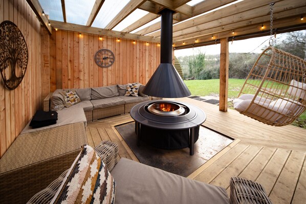 A cozy outdoor seating area with a wood-burning stove, a sofa, and a hanging chair.