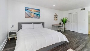 A bedroom with a large bed, a desk, a chair, a plant, and a painting on the wall. - Relaxation Starts Here (Zephyrhills)