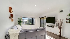 A modern living room with a grey sofa, a flat-screen TV, and a large window offering a view of greenery. - Relaxation Starts Here (Zephyrhills)
