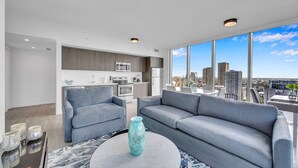 Een moderne woonkamer met een grijze bank, een ronde salontafel en een uitzicht op een stadsgezicht door grote ramen.