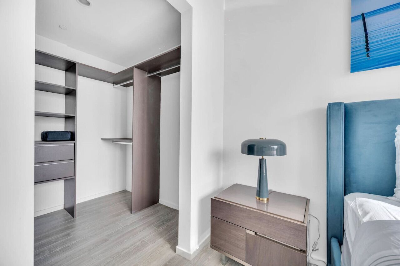 A modern hotel room with a bed, a bedside table with a lamp, a wardrobe, and a blue artwork on the wall.