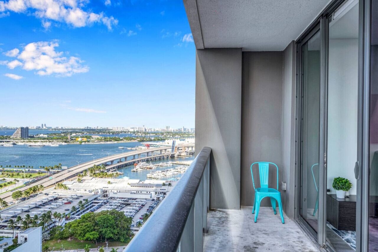 Un balcon avec une chaise turquoise, un pot de fleurs et une vue sur un port de plaisance et le paysage urbain.