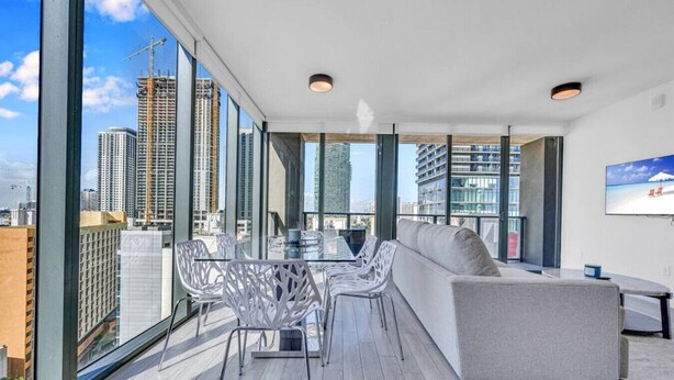 Eine moderne Dachterrasse mit Glastisch, weißen Stühlen, einer grauen Couch und einem Fernseher an der Wand.