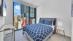 Ein modernes Schlafzimmer mit einem großen Bett, einem Schreibtisch und Blick auf eine Skyline durch ein großes Fenster.