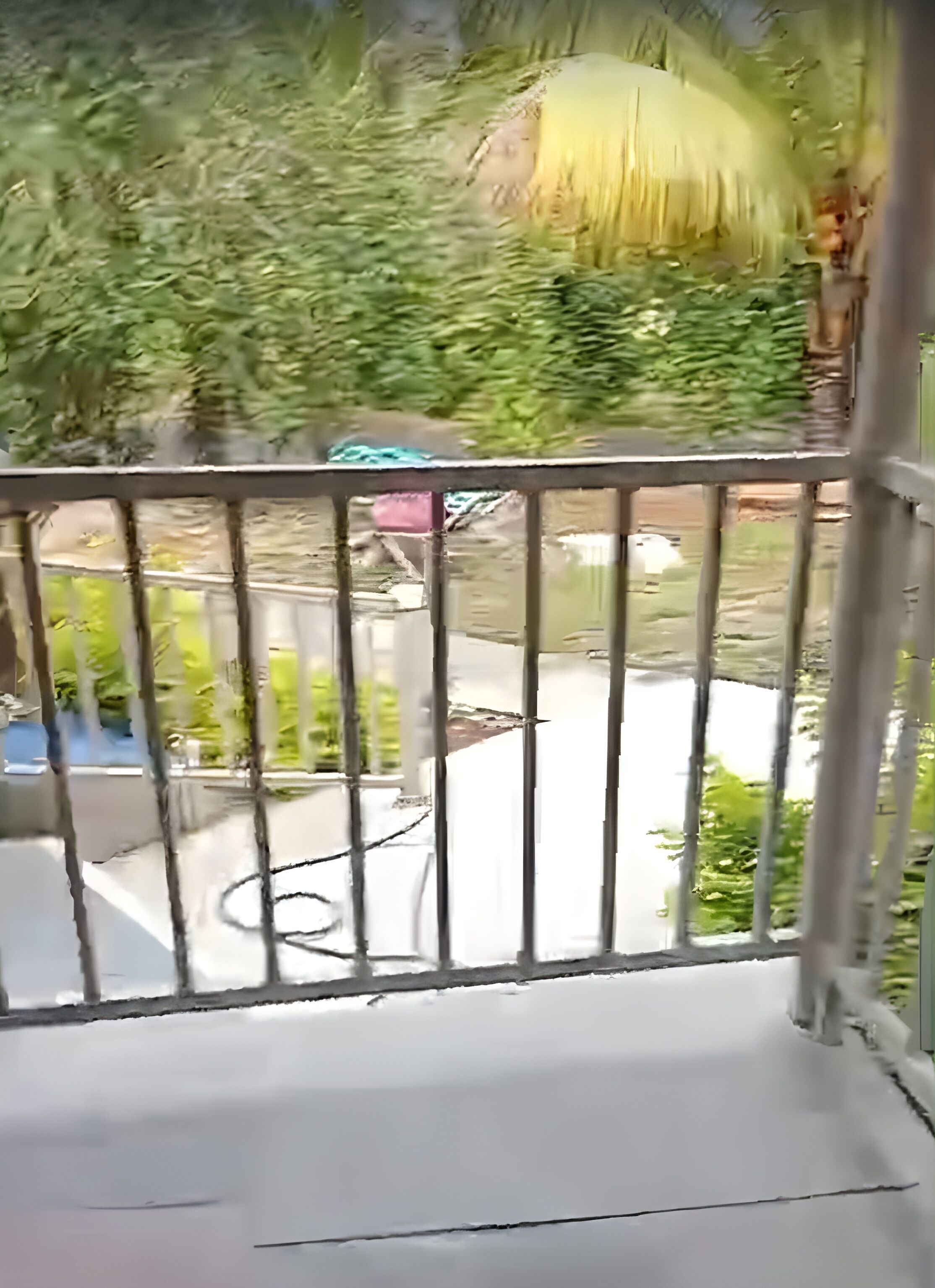 A balcony with a metal railing, a concrete floor, and a view of greenery and a building in the background.