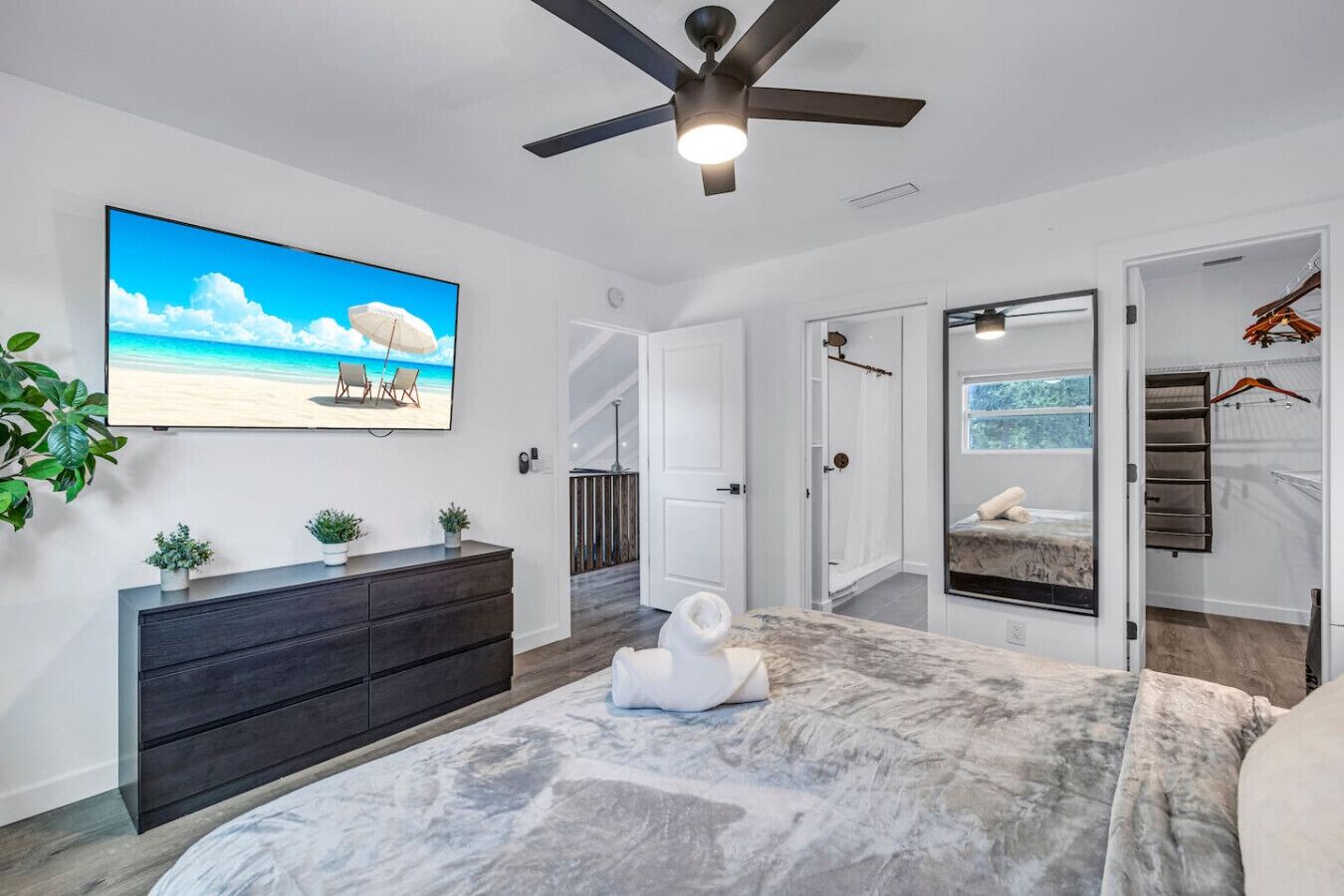 Ein modernes Schlafzimmer mit einem großen Bett, Deckenventilator, Fernseher der eine Strandansicht zeigt, begehbarem Kleiderschrank und Blick auf einen Pool.
