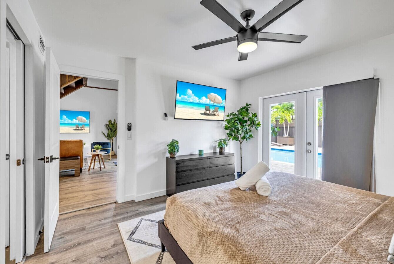 A bedroom with a bed, a ceiling fan, a TV, a dresser, and a view of the outside.