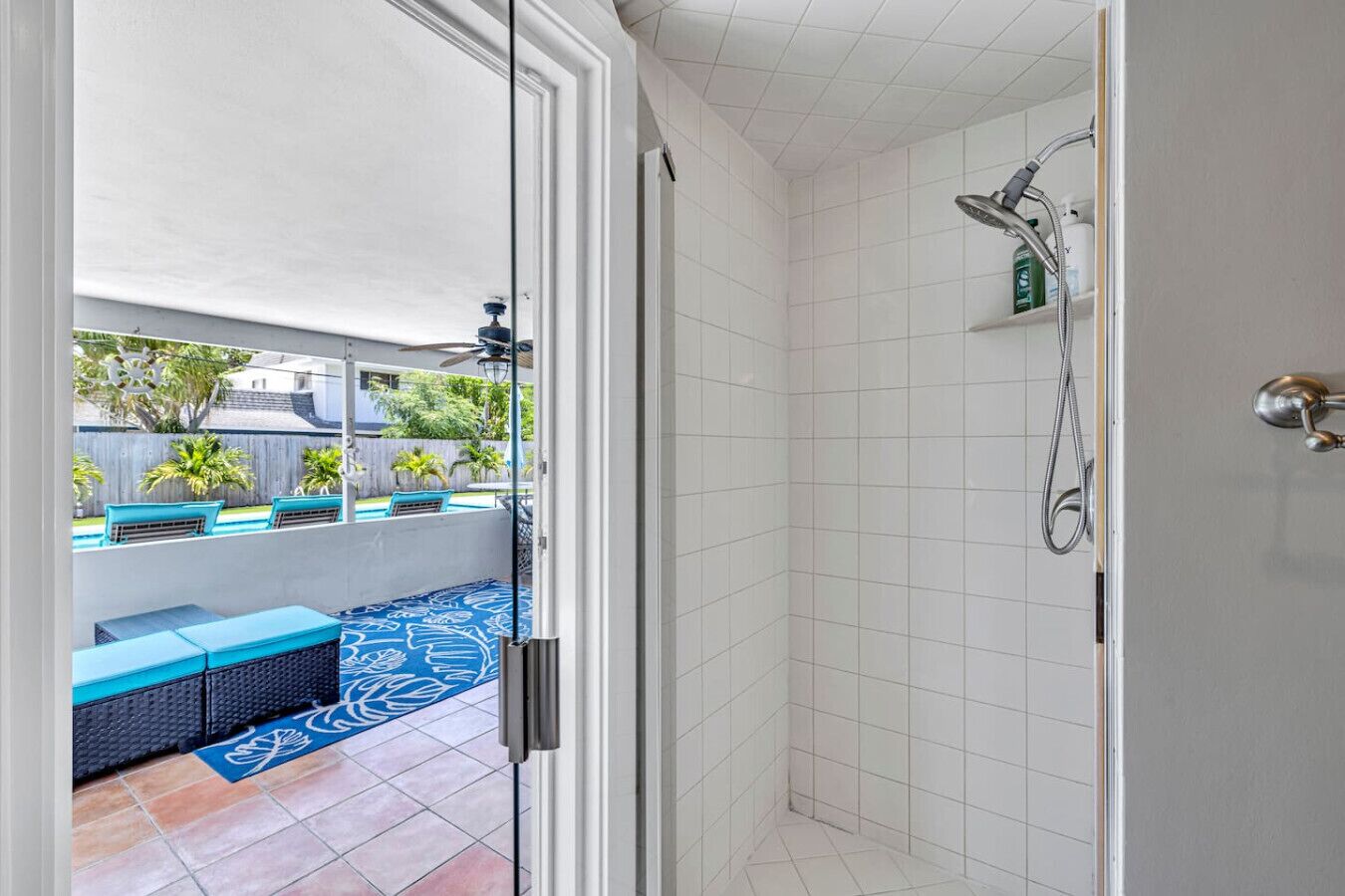 Un baño con ducha, un ventanal de vidrio que da a un patio con mobiliario exterior azul y una piscina.