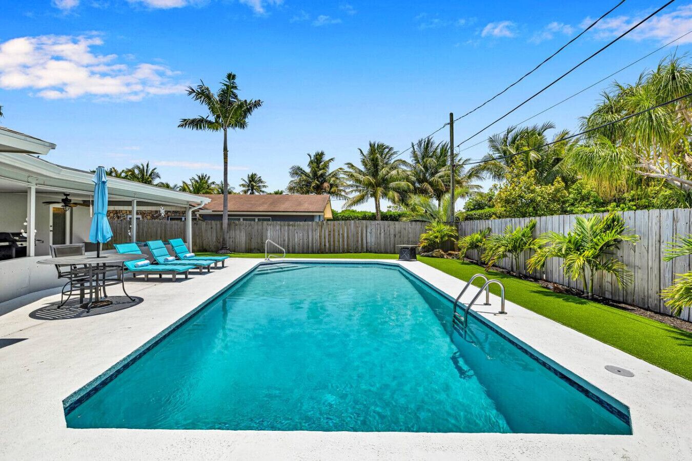 A rectangular swimming pool with a clear blue water, surrounded by a white deck. There are lounge chairs and a table with a blue umbrella.
