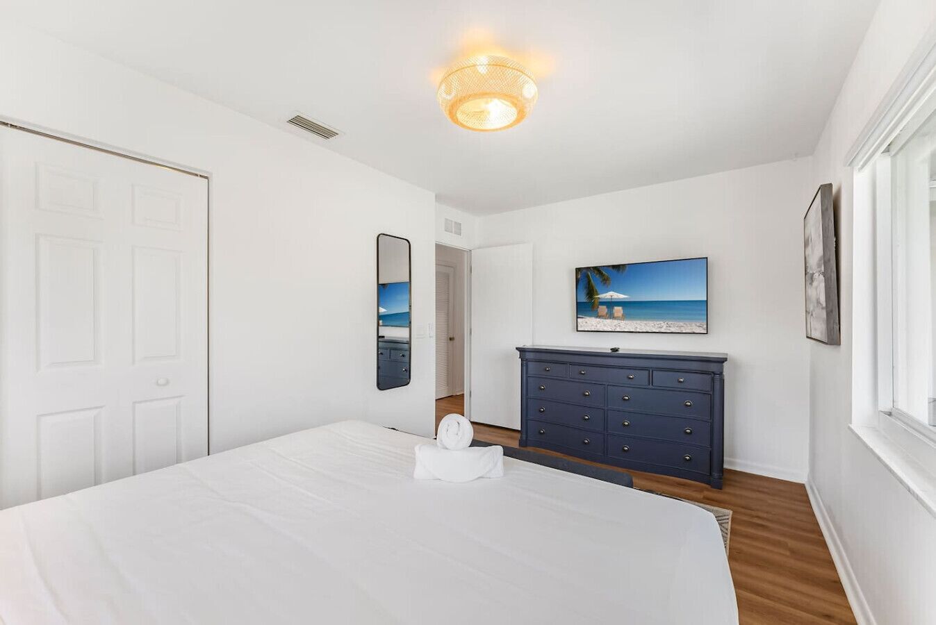 Une chambre à coucher avec un grand lit, une commode, un miroir, une télévision et un tableau représentant une scène de plage.