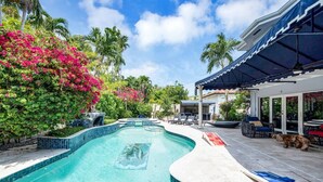 Ein Pool mit gewundener Kante, umgeben von üppiger Vegetation und einer überdachten Sitzgelegenheit mit Liegestühlen.
